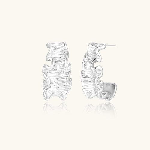 Textured white gold-plated hoop earrings with flowing wave pattern against cream background, showing dimensional rippled surface.
